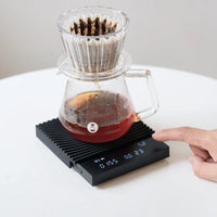 Timemore Black Mirror Basic 3.0 scale displaying live brew ratio while a V60 pour over drips into a glass server
