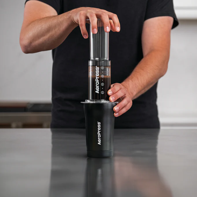 Person in a black t-shirt brewing coffee with an AeroPress Go Plus, pressing the plunger over a black AeroPress cup on a kitchen countertop, with a blurred background.