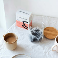 A box of Good & Proper English Breakfast tea bags with some in a glass jar with wooden lid, and a brewed cup of tea.
