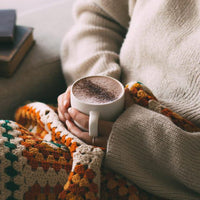 West Coast Cocoa Deluxe Hot Chocolate prepared in a mug with chocolate dusting on top, held by a person with a blanket