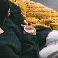 West Coast Cocoa Deluxe Hot Chocolate lifestyle shot prepared in a mug with chocolate dusting on top, held by a person with a blanket